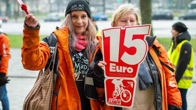 Zwei Frauen mit einem Schild auf dem "15 Euro" und das Liefern am Limit Logo zu sehen sind. 
