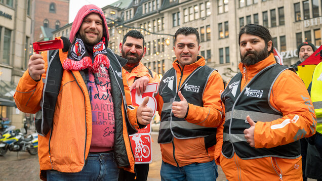 Vier Lieferando-Rider in orangenen Jacken stehen auf dem Rathausmarkt. 