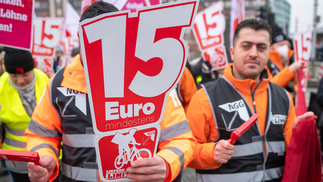Ein Mann in orangener Jacke hält ein Schild mit 15 Euro und dem Liefern am Limit Logo in die Kamera.
