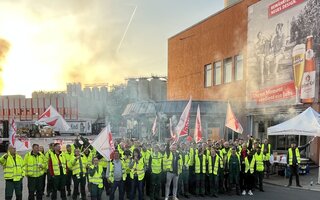 Große Gruppe Streikender in neongelben Warnwesten mit Fahnen und Pyrotechnik vor der Brauerei Berliner Kindl. 