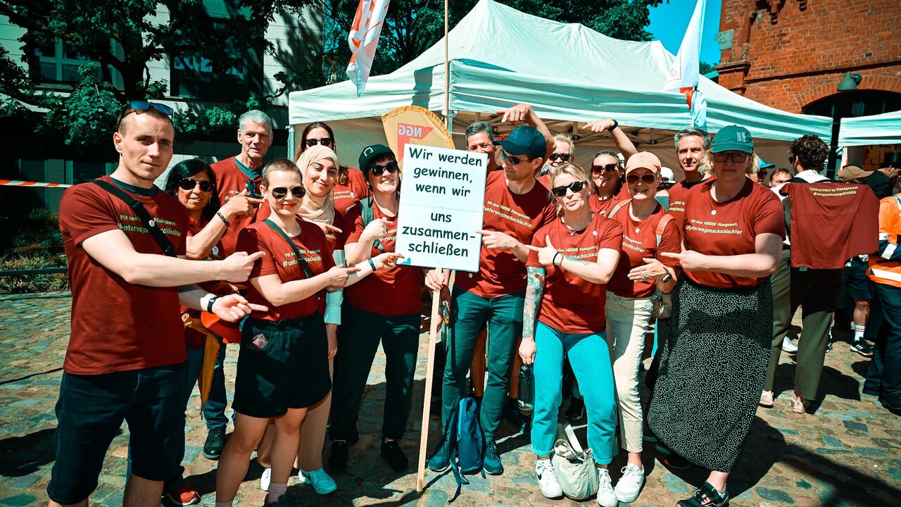 1_Mai_2025_Solifoto_Nestle_Hamburg Eine Gruppe Menschen steht im Halbkreis und hält in der Mitte ein Schild auf dem steht "Wir werden gewinnen, wenn wir und zusammenschließen."