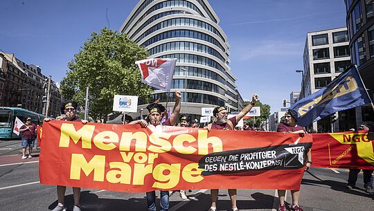Menschen halten ein großes Transparent auf dem "Mensch vor Marge" steht. Im Hintergrund sieht man die Nestlé-Zentrale in Frankfurt.