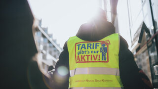 Ein Mann ist von hinten zu sehen. Er trägt eine neongelbe Warnweste auf der steht "Tarif gibt's nur aktiv!"