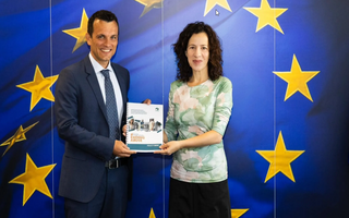 Ein Mann und eine Frau stehen vor einer großen Europa-Fahne und halten gemeinsam einen gedruckten Bericht in die Kamera. 