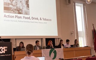 Ein Plenarsaal mit Menschen. Auf der Leinwand steht "Action Plan: Food, Drink, Tobacco"