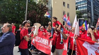 1_Mai_2025_Frankfurt_Demonstration Ein Mann hält ein Megafon währen ein kleines Kind auf den Schultern eines anderen Mannes sitzt und in das Megafon spricht. Alle befinden sich in einer großen Gruppe Menschen mit NGG-Fahnen.