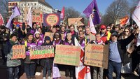 Eine Gruppe von Frauen und Männern posiert am 8. März für ein Gruppenfoto. Sie halten Schilder mit feministischen Sprüchen. 