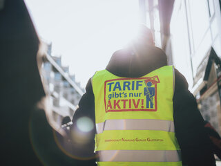 Das Bild zeigt einen Mann von hinten. Er trägt eine neongelbe Streikweste auf der "Tarif gibt's nur aktiv" steht.
