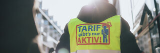 Das Bild zeigt einen Mann von hinten. Er trägt eine neongelbe Streikweste auf der "Tarif gibt's nur aktiv" steht.