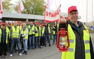 Im Vordergrund steht ein Mann mit einer gelben Streikweste und einer roten Laterne. Im Hintergrund stehen mehrere Menschen in gelben Streikwesten.