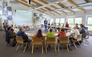 Eine Gruppe von ca. 20 Menschen sitz an Seminartischen in einem großen Raum und hört einer Referentin zu.