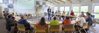 Eine Gruppe von ca. 20 Menschen sitz an Seminartischen in einem großen Raum und hört einer Referentin zu.