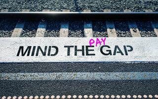 Auf dem Bild sieht man eine Bahnsteigkante auf der in weiß "Mind the Gap" steht. zwischen the und gap ist in pink das Wort "pay" eingefügt. 