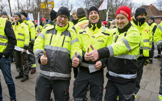 Drei Männer in neongelben Jacken lachen in die Kamera und strecken den Daumen nach oben als positives Zeichen. Im Hintergrund stehen noch mehr Männer in neongelben Jacken. Ein Mann trägt eine rote Mütze auf der "NGG" steht. 