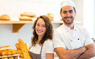 Eine junge Frau mit lockigen, braunen Haaren schaut seitlich in die Kamera. Sie steht Rücken an Rücken mit einem jungen Mann mit braunen, kurzen Haaren. Beide Tragen weiße Kleidung. Die Frau hält ein Baguette in der Hand und im Hintergrund sind Backwaren zu sehen. 