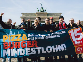 Mehere Mitglieder der Tarifkommission Systemgastronomie stehen hinter einem Banner auf dem "Pizza, Burger, Mindestlohn" steht. Sie schauen entschlossen und kämpferisch in die Kamera. 