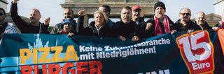 Mehere Mitglieder der Tarifkommission Systemgastronomie stehen hinter einem Banner auf dem "Pizza, Burger, Mindestlohn" steht. Sie schauen entschlossen und kämpferisch in die Kamera. 