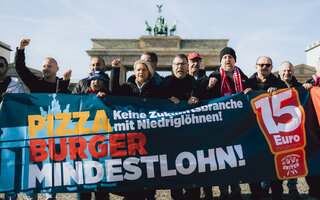 Mehere Mitglieder der Tarifkommission Systemgastronomie stehen hinter einem Banner auf dem "Pizza, Burger, Mindestlohn" steht. Sie schauen entschlossen und kämpferisch in die Kamera. 