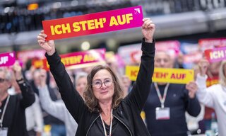 Eine Frau steht in einem Raum voll mit Menschen und hält ein Schild in die Luft auf dem "Ich stehe auf" steht.
