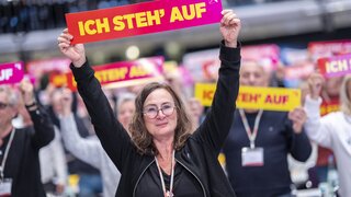Eine Frau steht in einem Raum voll mit Menschen und hält ein Schild in die Luft auf dem "Ich stehe auf" steht.