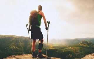 Ein Mann mit einer Schiene am Unterschenkel und Krücken steht auf einem Berg und schaut in die Ferne.