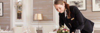 Eine Frau in einem dunkelblauen Kostüm richtet einen Tisch in einem Hotelrestaurant her. Der Tisch ist fein gedeckt mit einer weißen Tischdecke und Blumen. 