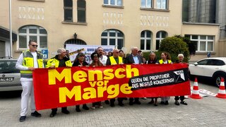 Menschen stehen hinter einem großen Banner auf dem "Mensch vor Marge" steht. 