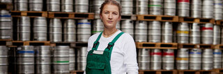 Eine junge, blonde Frau in grüner Arbeitslatzhose steht vor einer Wand aus Bierfässern. Sie schaut direkt und aufrecht in die Kamera.