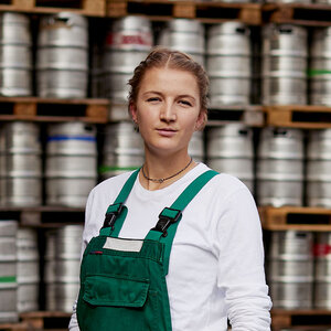 Eine junge, blonde Frau in grüner Arbeitslatzhose steht vor einer Wand aus Bierfässern. Sie schaut direkt und aufrecht in die Kamera. 