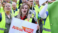 Zwei junge Frauen mit einer Fahne von jungeNGG und neongelben Streikwesten stehen im Vordergrund. Im Hintergrund sieht man eine Menge mit Streikwesten. 