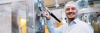 Ein Mann in weißer Schutzkleidung steht an einer Maschine. Er wendet sich der Kamera zu und lächelt.