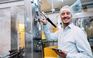 Ein Mann in weißer Schutzkleidung steht an einer Maschine. Er wendet sich der Kamera zu und lächelt. 