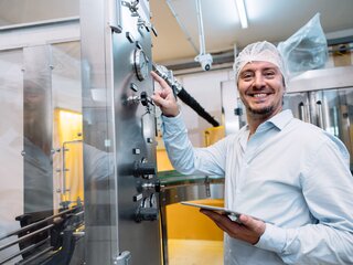 Ein Mann in weißer Schutzkleidung steht an einer Maschine. Er wendet sich der Kamera zu und lächelt.