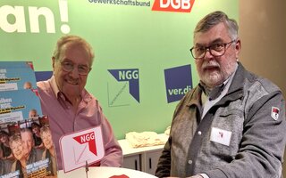Zwei ältere Männer stehen an einem Stehtisch und schauen in die Kamera. 