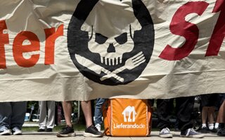 Ein orangener Lieferrucksack von Lieferando steht auf dem Boden unter einem großen Transparent auf dem Lieferstreik steht und ein Totenkopf zu sehen ist.